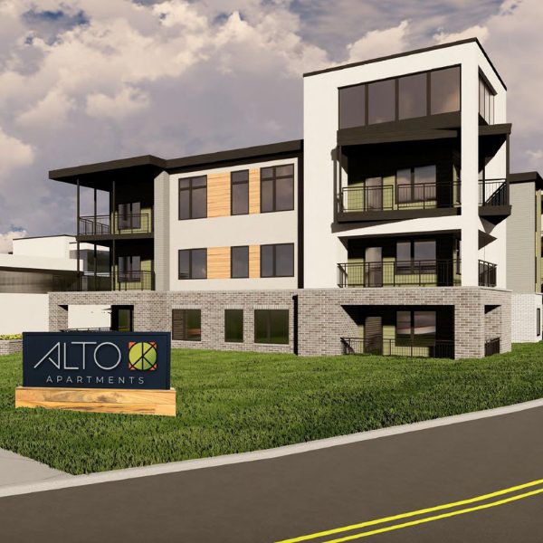 Alto Apartments Residential Buildings A modern living room with a loft design features a metal staircase leading to an upper level. Two large windows with blinds allow natural light to fill the space. The room includes a gray couch, a coffee table with a glass fireplace, and a floor lamp.